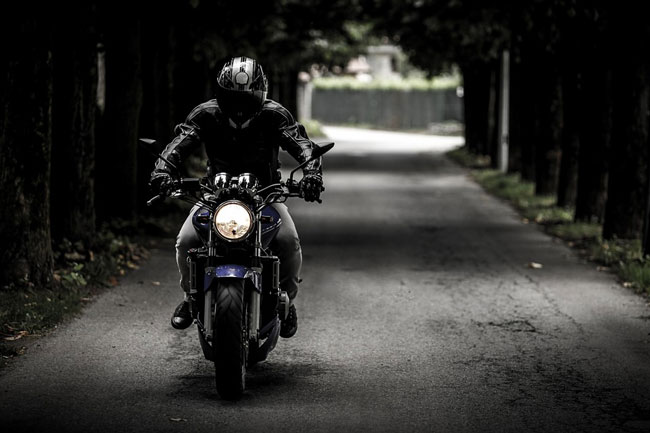 A motorcyclist dressed all in black rides through an alleyway completely shaded by trees.