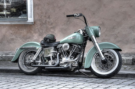 A forest green vintage Harley-Davidson Low Rider from 1977 sits parked on the curb of a gray brick road.